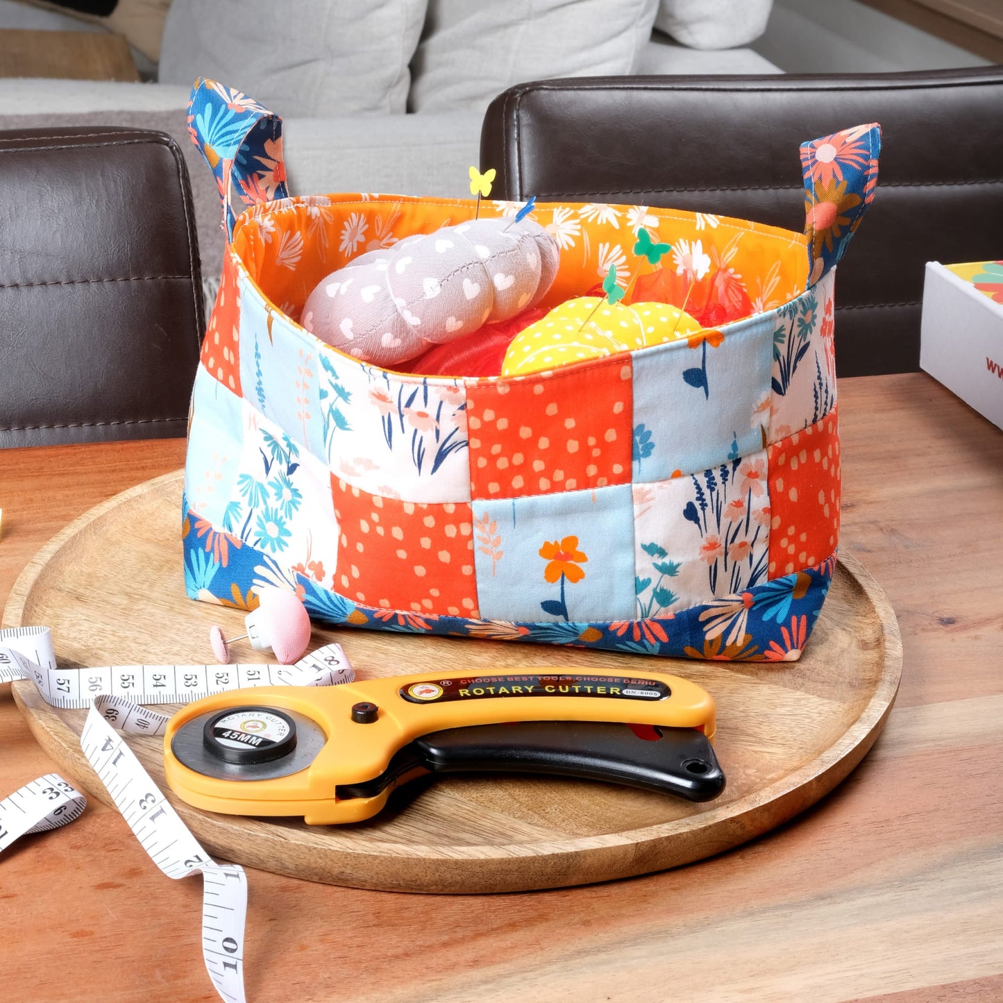 The Mrs Quilty Subscription Box features a vibrant fabric basket with stuffed vegetables on a round wooden tray. Nearby, you'll find a yellow rotary cutter and measuring tape on the wooden table.