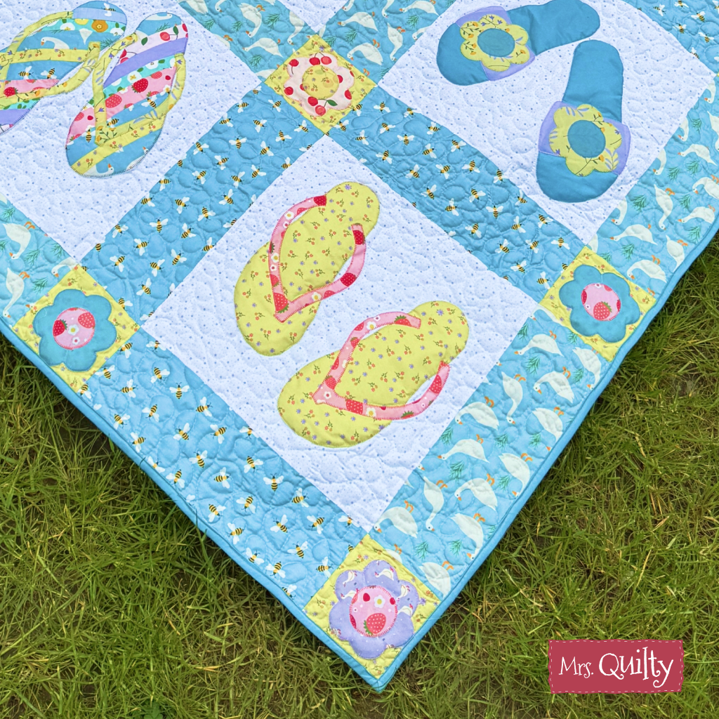 The Flip Flop Quilt Downloadable PDF Quilt Pattern features a playful design with appliqué flip-flops and flowers on green grass, finished with a “Mrs. Quilty” label in the corner.