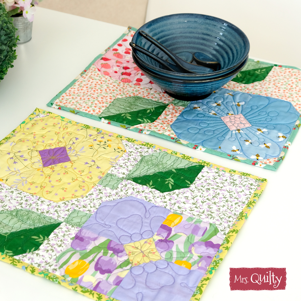 Two colorful quilted placemats with floral designs, made from the "Flower Placemats Downloadable PDF Quilt Pattern," are shown with a blue ceramic bowl and black chopsticks on a white surface; "Mrs. Quilty" label appears in the corner.