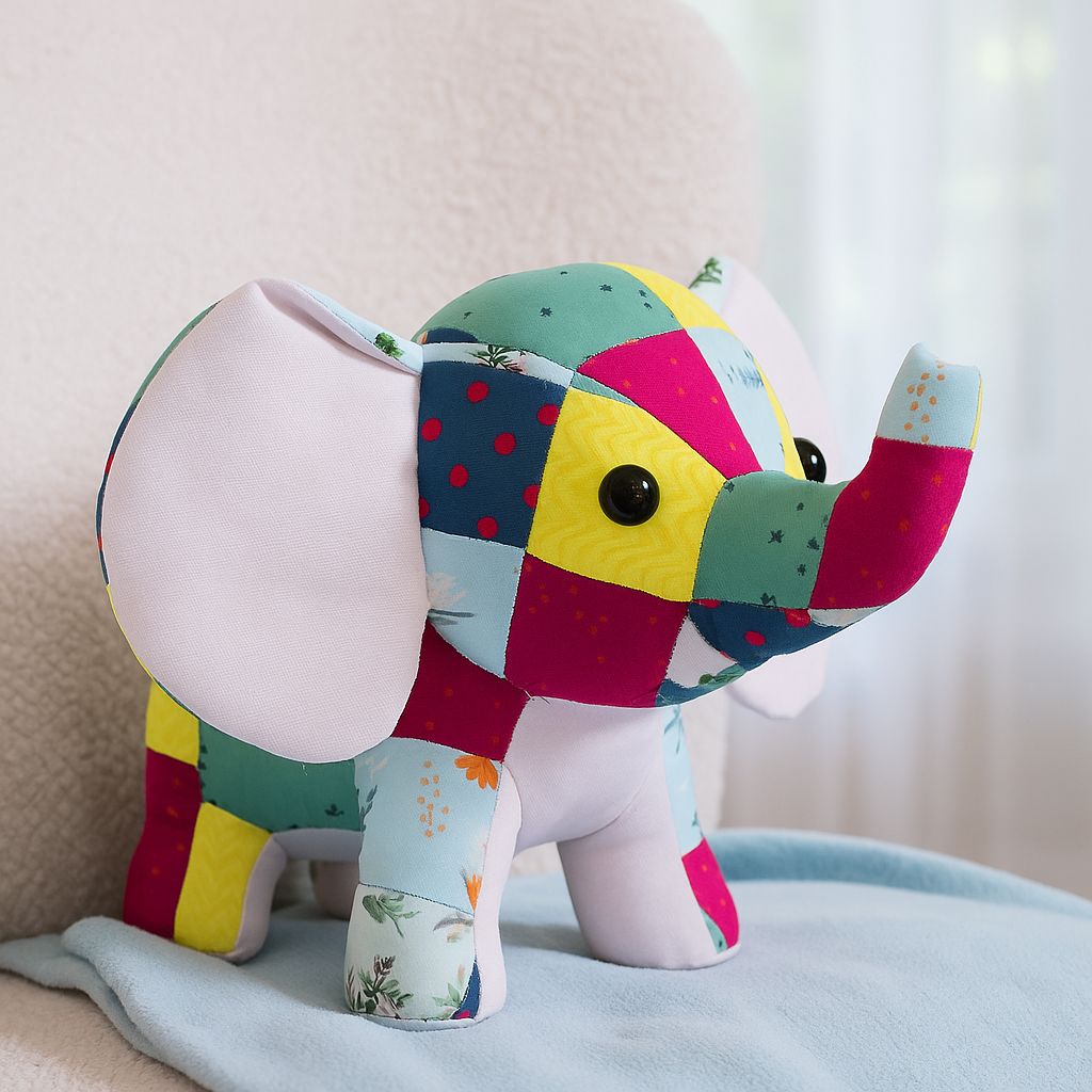 A softie made from the Quilo the Elephant Downloadable PDF Quilt Pattern rests on a light blue blanket, its vibrant patchwork bright—making it a perfect handmade sewing gift for any occasion.