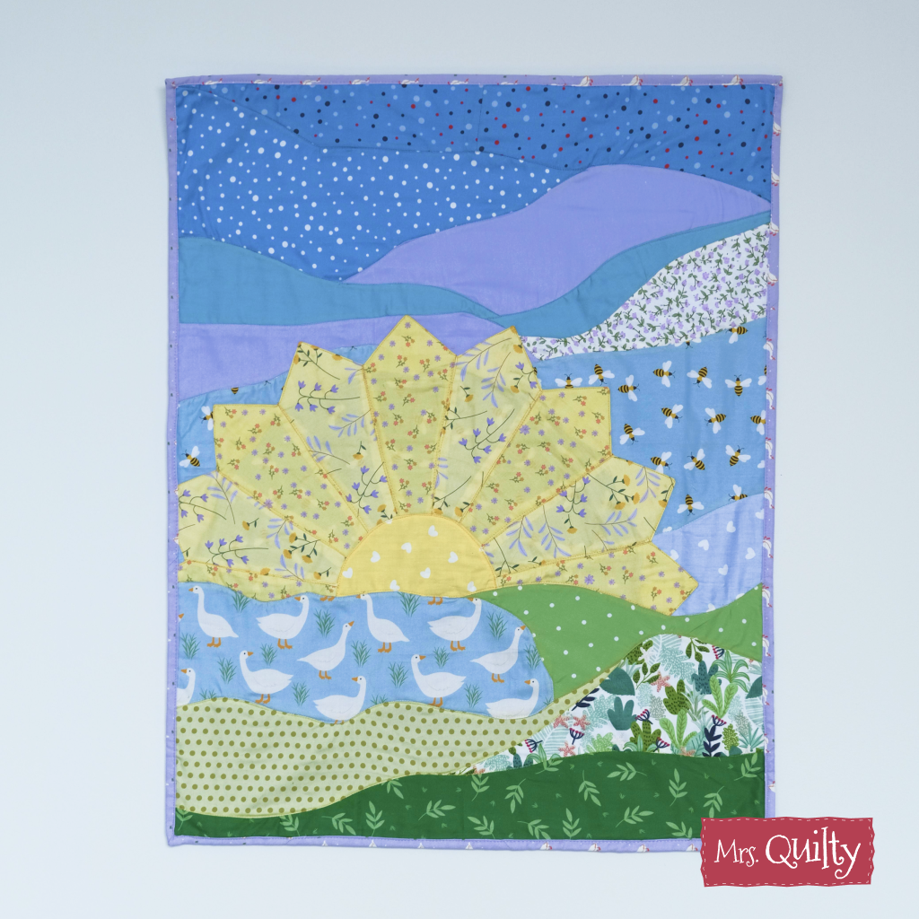 The Sunrise Wall Hanging Downloadable PDF Quilt Pattern features a vibrant quilt design with a yellow sunburst, blue mountains, and green hills in various patterns, shown on a plain white background.