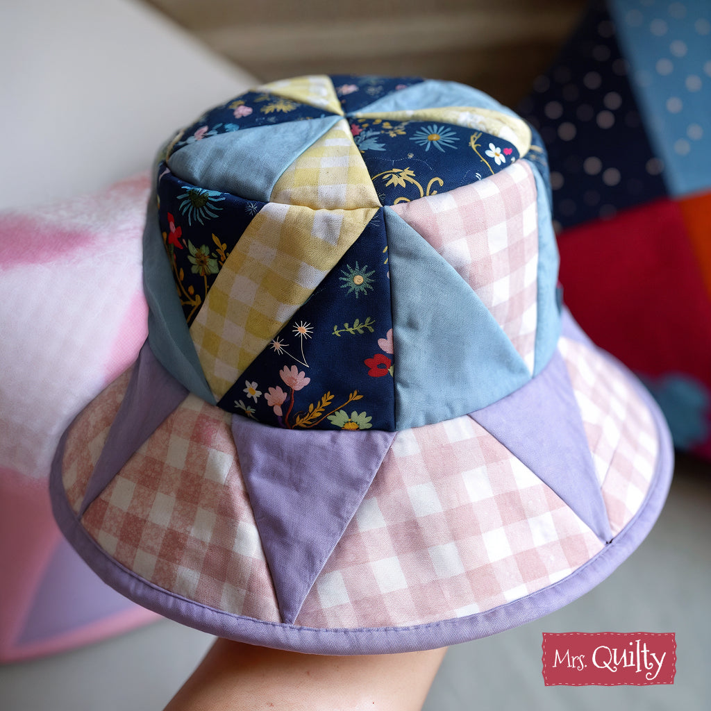 Held in hand indoors, the "Mrs. Quilty" logo appears in the corner of a patchwork bucket hat made with pastel and floral panels using the Bucket Hat Downloadable PDF Quilt Pattern.