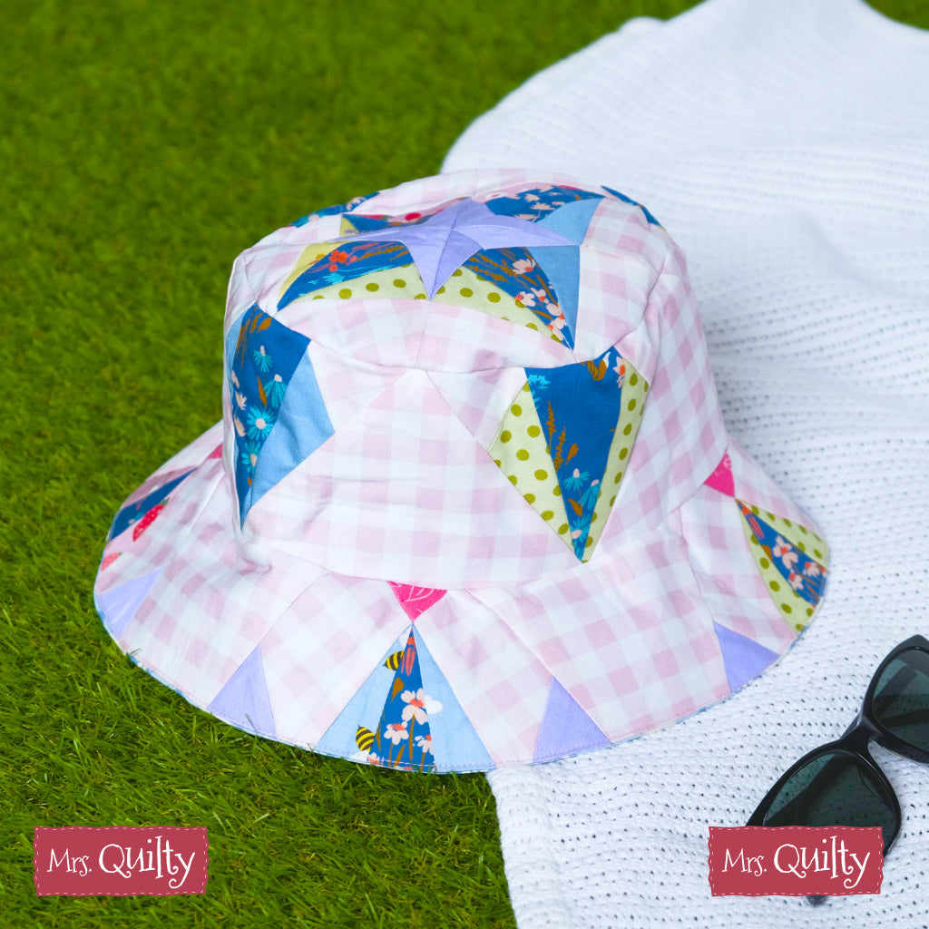 The Bucket Hat Downloadable PDF Quilt Pattern features an easy quilting design with pastel and floral triangles, displayed on white fabric next to black sunglasses on green grass.
