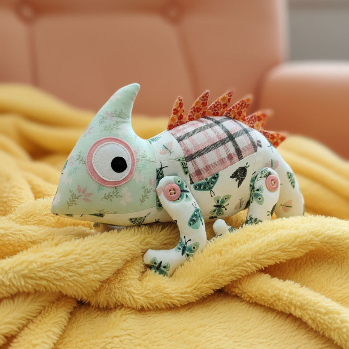 Chichi the Chameleon Downloadable PDF Quilt Pattern features an advanced quilting design to create a handmade stuffed chameleon toy, perfect for displaying on a yellow blanket with a peach-colored sofa in the background.