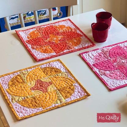 Three vibrant quilted "Blazing Blooms" mug rugs from the downloadable PDF pattern are shown on a white table with two red cups. The "Mrs. Quilty" logo appears in the bottom right corner.