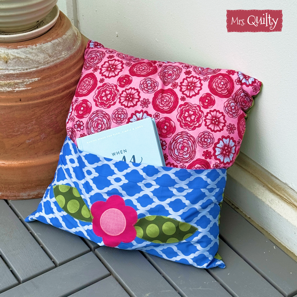 A Reading Pillow Downloadable PDF Quilt Pattern features appliqué details and a blue book pocket, shown on a gray deck next to a large terracotta pot.
