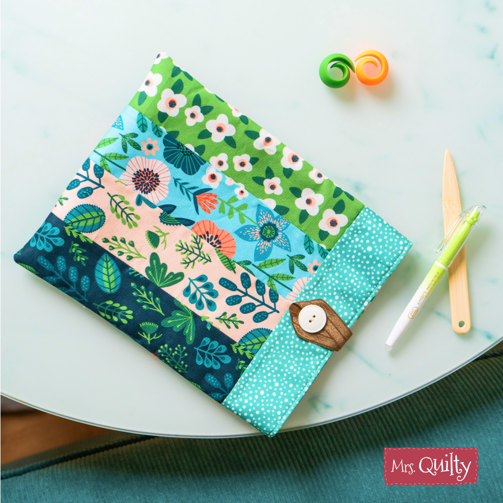 A Book Sleeve Downloadable PDF Quilt Pattern featuring colorful floral and leaf designs, button closure, and accessory pockets is displayed on a round table with the Mrs. Quilty logo in the lower right corner.
