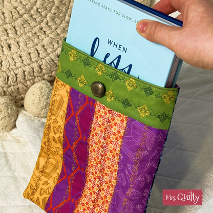 A hand slides a blue book into a vibrant, striped quilted cover with a snap button and "Mrs. Quilty" tag. Made using the Book Sleeve Downloadable PDF Quilt Pattern, this padded sleeve protects your book in style on any surface.