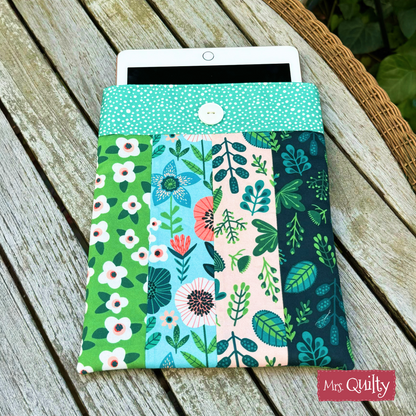 A tablet partially slides out of a padded book sleeve made with the Book Sleeve Downloadable PDF Quilt Pattern, featuring colorful floral and leaf designs, resting on a weathered wooden table.