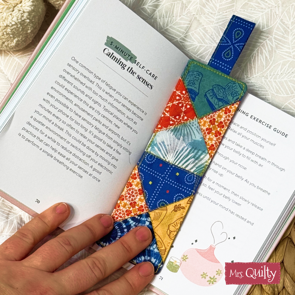 A hand holds open a book to a self-care page, featuring a colorful, quilted fabric bookmark made from our Bookmark Downloadable PDF Quilt Pattern—the perfect inspiration for your next instant digital craft project.