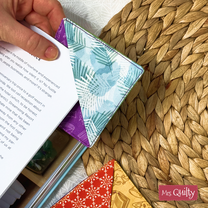 A hand places a book with a blue and purple corner bookmark made from the Corner Bookmark Downloadable PDF Quilt Pattern on a woven mat. Another book with a red and yellow bookmark using the same pattern rests nearby.