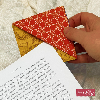 A hand places a red and yellow corner bookmark, made using the Corner Bookmark Downloadable PDF Quilt Pattern, on a book page. "Mrs. Quilty" appears in the corner.
