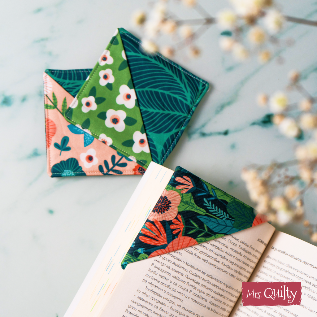 Two colorful, triangular bookmarks made from quilting fabric scraps with floral and leaf patterns demonstrate the Corner Bookmark Downloadable PDF Quilt Pattern; one marks a page in an open book on a marble surface.