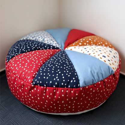 A round quilted floor cushion made with the Floor Cushion Downloadable PDF Quilt Pattern features colorful, patterned fabric sections in red, blue, orange, and white with small dotted designs, perfect for accenting rooms with dark flooring.