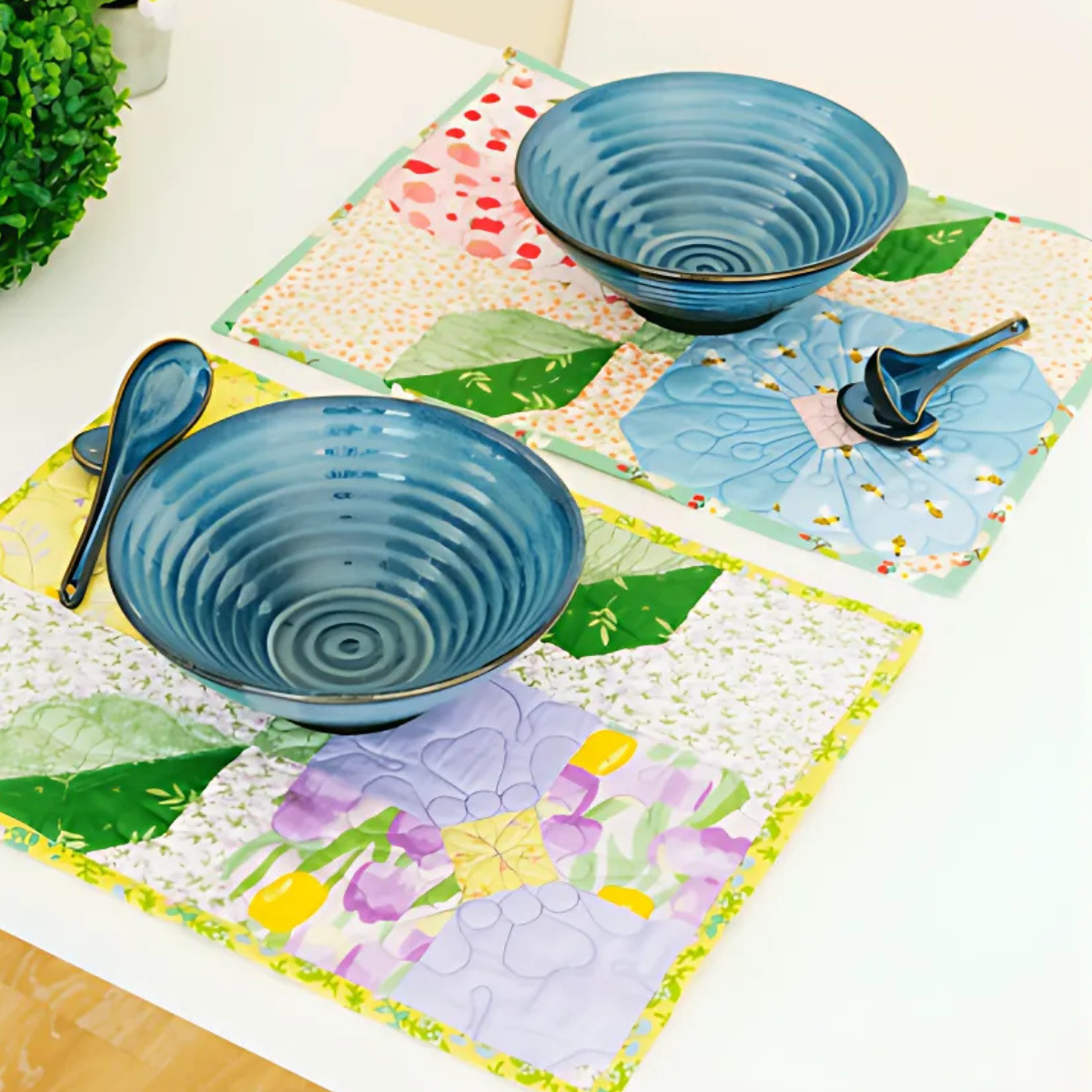Two place settings with blue ceramic bowls, matching spoons, and chopsticks sit on a white table atop colorful placemats made from the Flower Placemats Downloadable PDF Quilt Pattern.