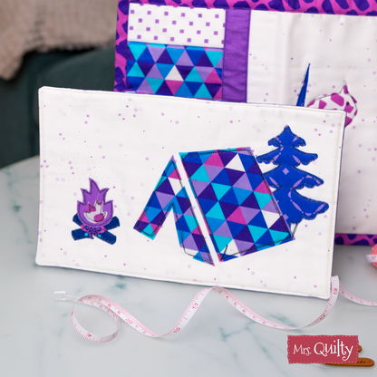 Featuring geometric designs of a tent, tree, and campfire in purple and blue, the Hiking Mug Rug Downloadable PDF Quilt Pattern is ideal for creating a unique mug rug with a playful outdoor theme.