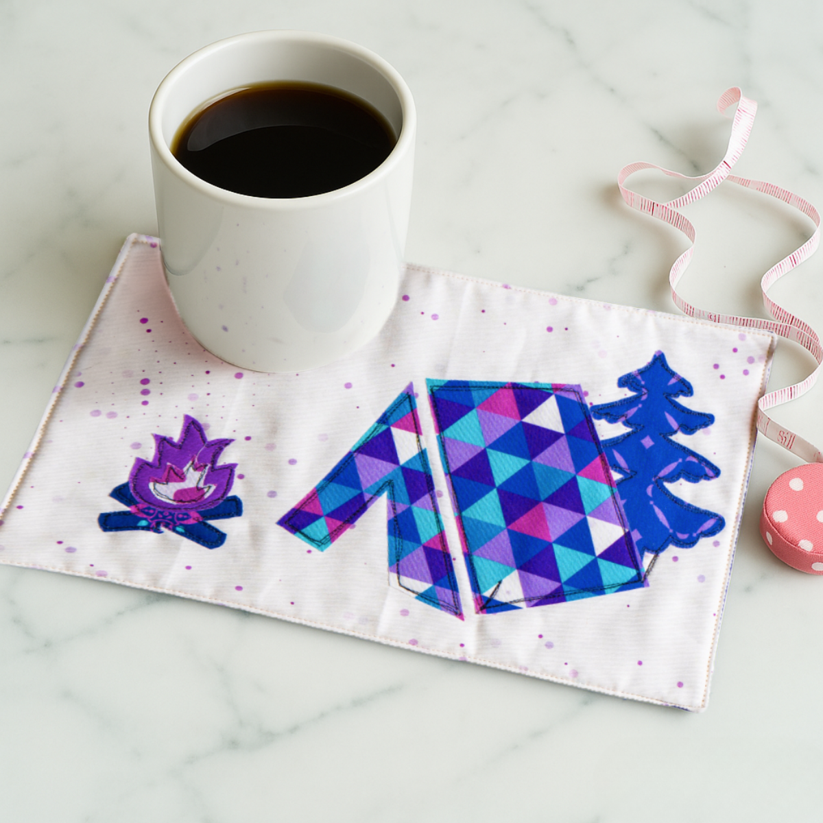 A cup of black coffee rests on a quilted rectangular cloth made with the Hiking Mug Rug Downloadable PDF Quilt Pattern, featuring colorful camping images—ideal for outdoor lovers. A pink measuring tape and button are nearby on the marble surface.