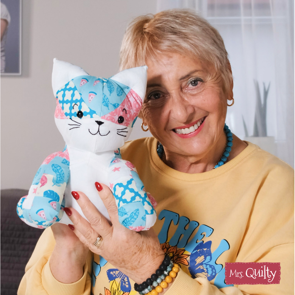 A smiling woman sits indoors holding a colorful quilted cat toy made from the "Katie the Kitten Downloadable PDF Quilt Pattern," showing off her quilting skills. The text "Mrs. Quilty" appears in the lower right corner.