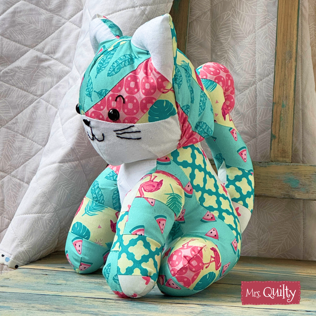 A vibrant quilted cat toy, crafted from teal, pink, and yellow fabrics with fruity leaf motifs, sits on wood. The label "Mrs. Quilty" appears in the corner. Pattern: Katie the Kitten Downloadable PDF Quilt Pattern.