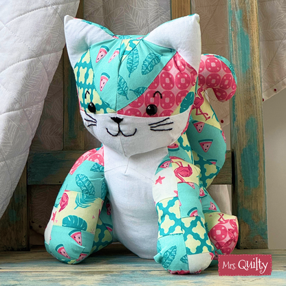 A Katie the Kitten downloadable PDF quilt pattern features a cute cat design with vibrant fabric patterns, perfect for showcasing your quilting skills. The pattern displays the cat sitting upright against a wooden backdrop.