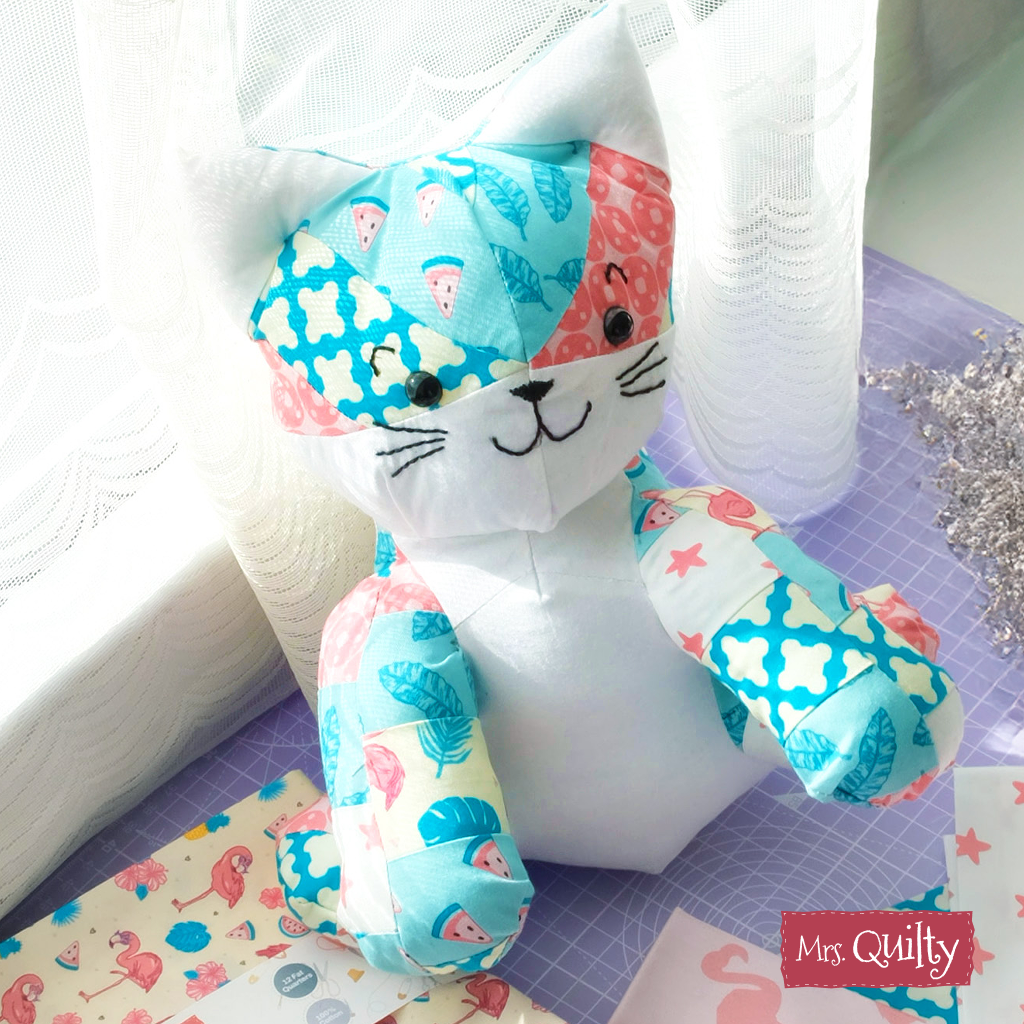 A "Katie the Kitten" quilt, made from blue, pink, and white fabrics using the downloadable PDF pattern, rests on a purple surface near a window with sewing materials and a "Mrs. Quilty" label nearby.