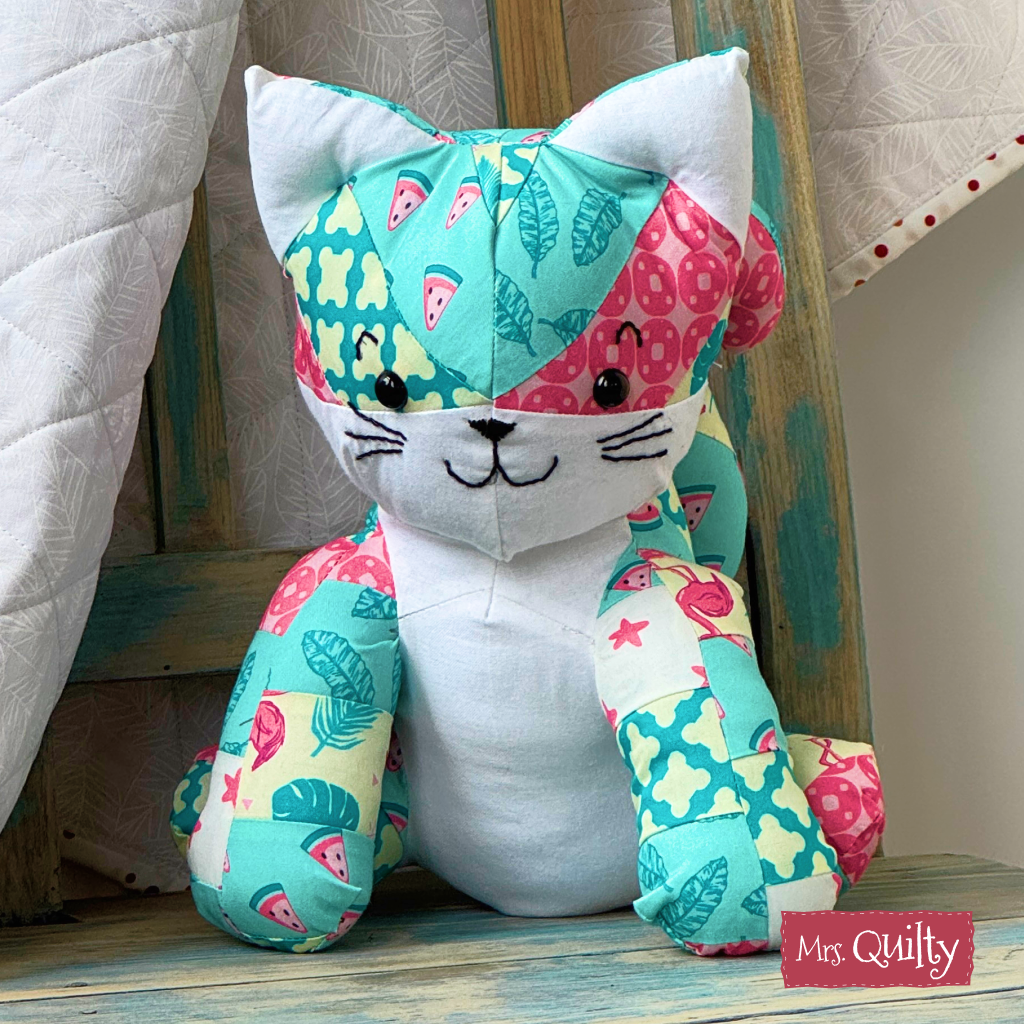 Katie the Kitten Downloadable PDF Quilt Pattern features a colorful patchwork cat design. Displayed on a wooden chair with a quilt in the background, the "Mrs. Quilty" logo is in the lower right corner.