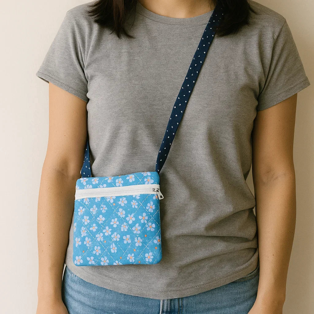 A person in a grey t-shirt and blue jeans carries a small blue floral bag made from the Quilted Crossbody Bag Downloadable PDF Quilt Pattern, featuring a dark blue strap and white zipper.