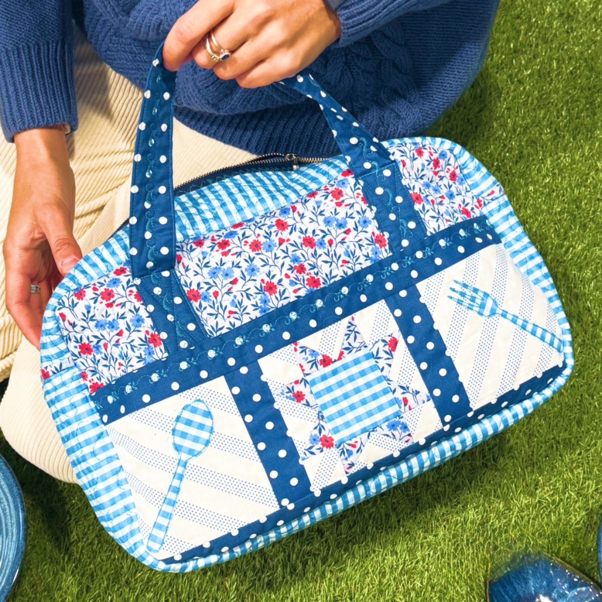 A person sits on green grass holding a blue and white lunch bag featuring floral, gingham, and polka dot designs made from the Quilter’s Lunch Bag Downloadable PDF Quilt Pattern.