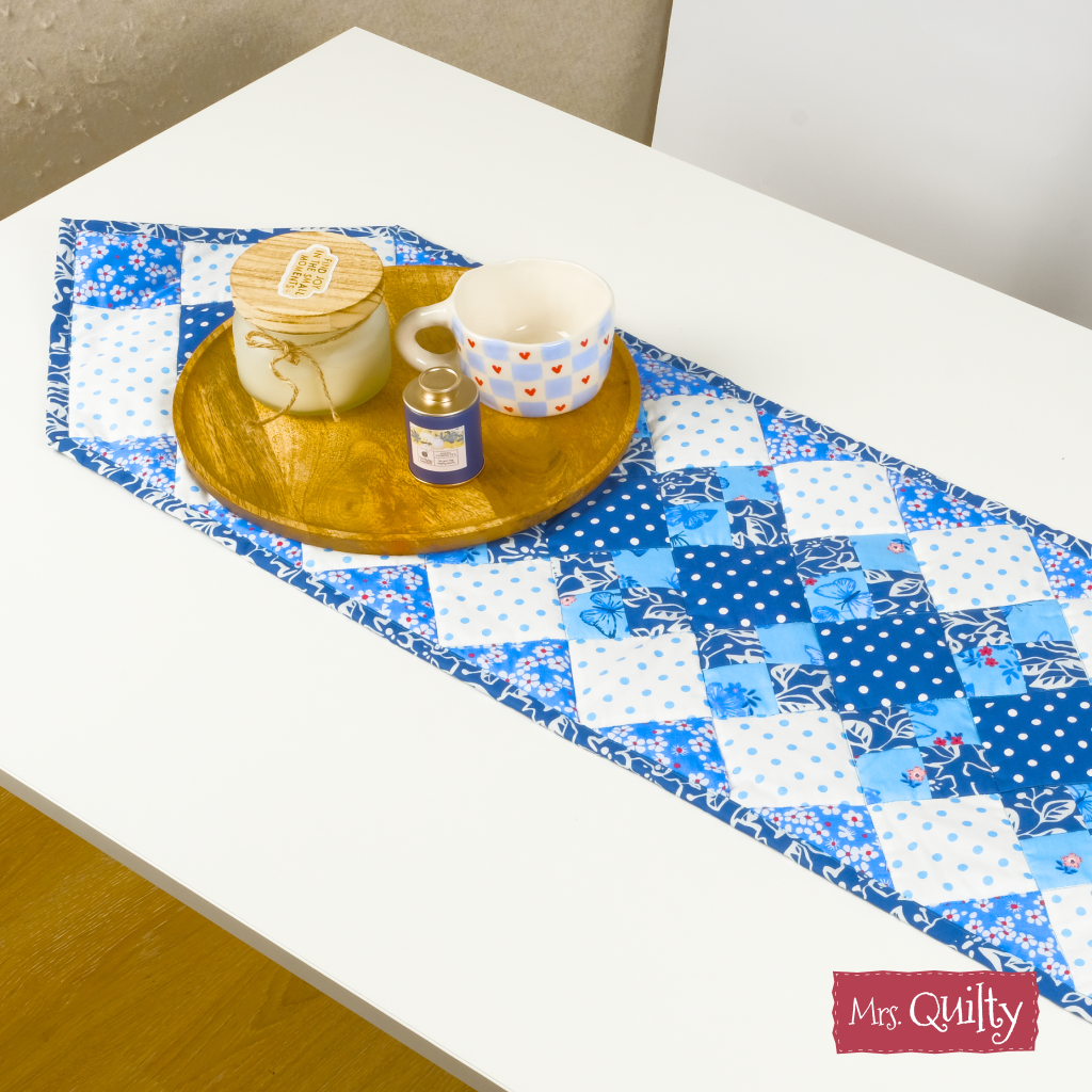 A wooden tray with a candle, jar, and white mug sits on a blue table runner made from the Table Runner Downloadable PDF Quilt Pattern atop a white table.
