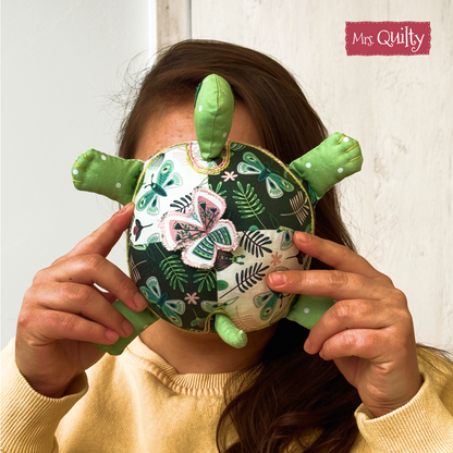 A person holds a plush quilted turtle toy in front of their face, obscuring their features. The toy is made using the "Timmy the Turtle Downloadable PDF Quilt Pattern," making it a charming choice for handmade gifts.