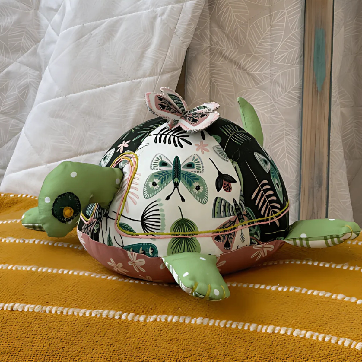 Timmy the Turtle Downloadable PDF Quilt Pattern features a fabric turtle with floral and butterfly details, perfect for beginners and easily displayed on a mustard yellow striped surface near a white quilted background.