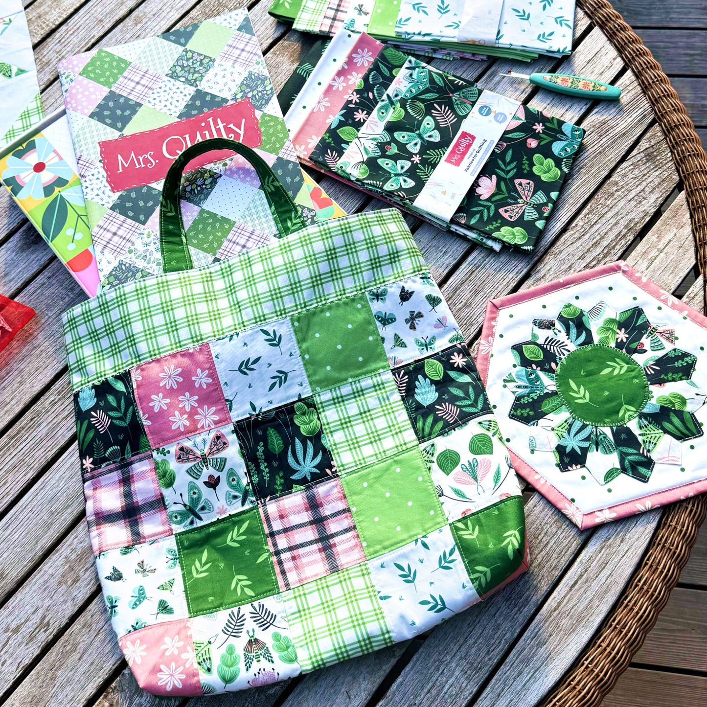 A wooden table displays a colorful patchwork tote with green and pink floral designs, complemented by matching fabrics, a quilted coaster, and the "Mrs Quilty Subscription Box" magazine.