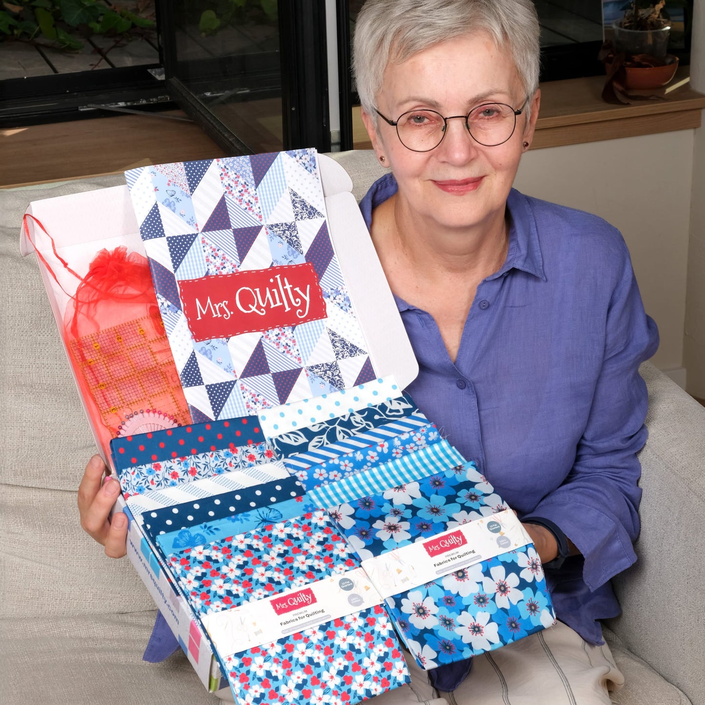 A person with short gray hair and glasses holds an open "Mrs Quilty Subscription Box" full of colorful quilt samples, seated on a light-colored couch.